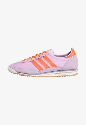 Niskoprofilowy sneaker Adidas w jasnofioletowym kolorze z pomarańczowymi paskami, pomarańczowymi sznurówkami i białą podeszwą z gumowym bieżnikiem.