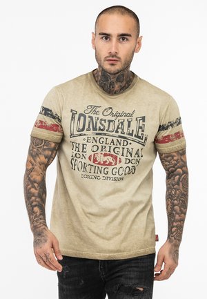 Tan cotton t-shirt with short sleeves, featuring a large printed logo and text in dark blue and red. Accented with striped fabric on sleeves.