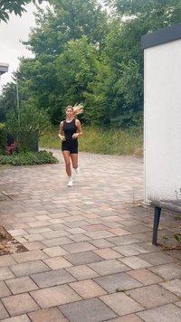 Joven corriendo por un camino pavimentado cerca de arbustos y árboles verdes, vistiendo un atuendo atlético negro y zapatillas blancas.