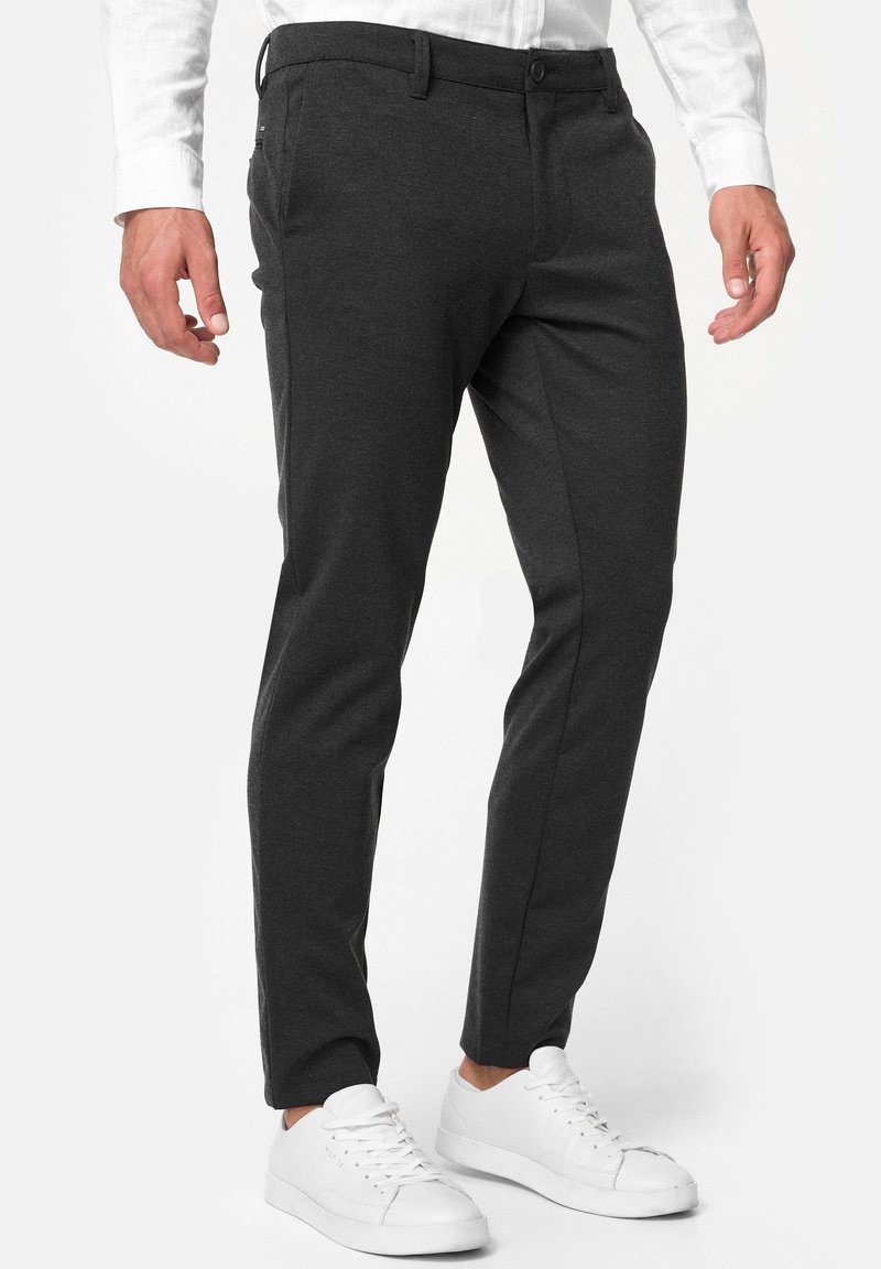 Homme portant un pantalon gris foncé ajusté, associé à des baskets blanches et une chemise blanche à manches longues, debout sur un fond clair.