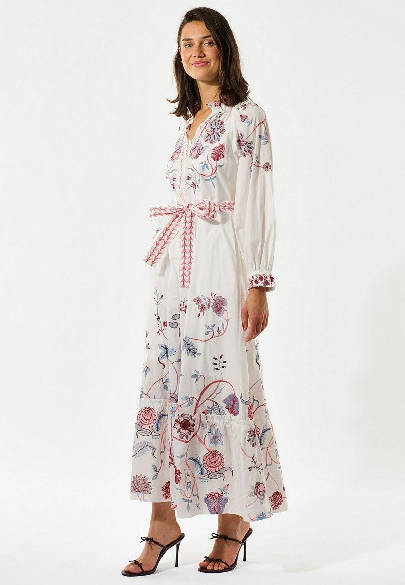 Femme portant une robe longue à manches longues blanche avec une broderie florale rouge et bleue, une ceinture à motif, et des sandales à talons hauts noires.