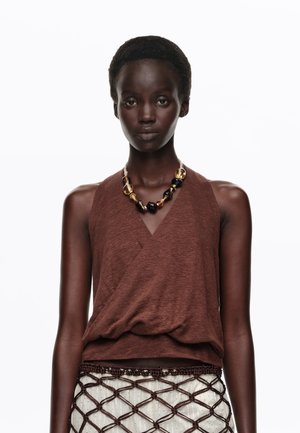 Mujer que lleva un top marrón sin mangas tipo wrap, un collar grueso de cuentas y una falda estampada con una superposición de red marrón.