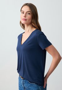 Granatowa koszulka V-neck w miękkim materiale, dopasowany fason, krótkie rękawy, z delikatnym drapowaniem przy dolnej krawędzi, zestawiona z jasnoniebieskimi dżinsami.