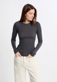 Donna con lunghi capelli castani, che indossa una maglietta aderente grigia scura a maniche lunghe e pantaloni beige chiaro, con le mani nelle tasche, mentre guarda verso il basso di lato.