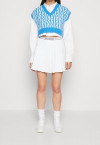 Ljust blå, stickad kort tröja med V-ringning och vita kabelmönster, matchad med en vit, plisserad kjol och vita sneakers med ankelsockor.