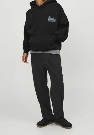 Sudadera negra con un bolsillo delantero, que presenta un pequeño gráfico multicolor en el pecho. Combinada con pantalones negros holgados y zapatos negros.