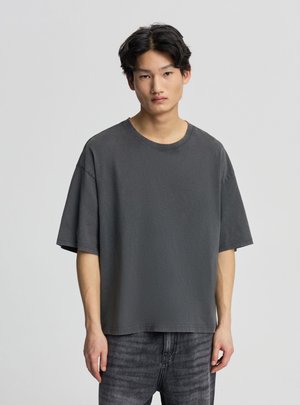 Giovane uomo con capelli scuri e mossi che indossa una maglietta larga grigio scuro e jeans neri sbiaditi su uno sfondo chiaro e uniforme.