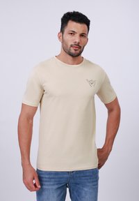Beige cotton t-shirt with short sleeves, round neck, and subtle shaka hand gesture graphic on left chest. Classic fit, smooth texture.