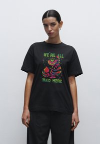 Zwarte katoenen t-shirt met kleurrijk grafisch ontwerp met de tekst "WE ZIJN ALLEMAAL GEK HIER" en speelse karakterillustraties.