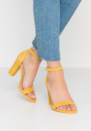 Voeten met gele suède sandalen met hoge hakken en enkelbandjes, gecombineerd met lichtblauwe jeans met rafelige zoom.