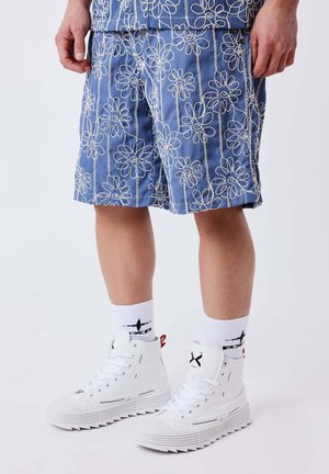 Mann trägt blaue Shorts mit Blumenmuster, weiße High-Top-Sneaker mit schwarzen Akzenten und weiße Crew-Socken mit schwarzem Design.