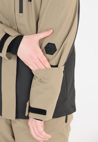 Giacca esterna beige e nera con logo esagonale testurizzato, polsini regolabili con Velcro e tasca con zip sulla manica.