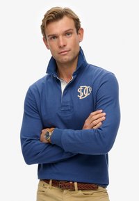 Blue long-sleeve polo shirt with standing collar, featuring a light-colored placket and embroidered logo on the chest. Worn with khaki pants.
