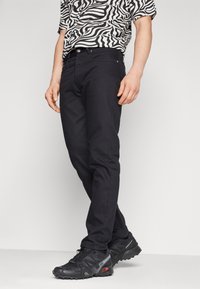 Jeans de mezclilla negra con corte ajustado, que presentan un diseño clásico de cinco bolsillos y costuras sutiles. Llevado con zapatillas deportivas negras.