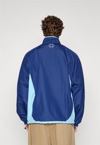 Μπουφάν αδιάβροχο σε ναυτικό μπλε χρώμα με ανοιχτό μπλε λεπτομέρειες στα πλαϊνά και ψηλό γιακά. Διαθέτει ελαστικά μανίκια και ρυθμιζόμενο κάτω μέρος με κορδόνι. Λογότυπο στην πλάτη.