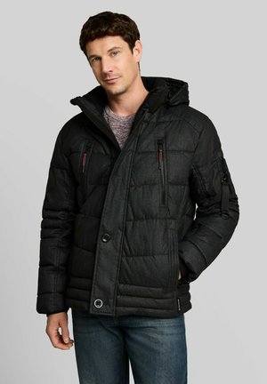Uomo con giacca invernale trapuntata nera con tasche sul petto con cerniera e cappuccio, abbinata a jeans blu, su sfondo grigio semplice.