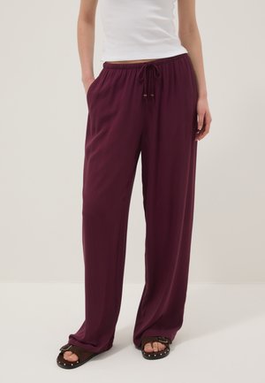 Persona con pantalones burdeos sueltos de cintura alta con cordón y sandalias marrones, de pie frente a un fondo claro y liso.