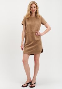 Blondynka pozuje w krótkim rękawie, sukience z zamszu w kolorze beżowym sięgającej do kolan oraz czarnych sandałach na obcasie na tle jednolitego białego tła.