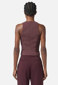 Top fitted senza maniche color borgogna con collo alto, tessuto testurizzato e dettagli con zip laterali, abbinato a pantaloni su misura coordinati.