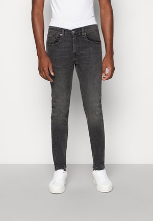 Mand iført sorte slim-fit jeans, hvide sneakers og en hvid tætsiddende t-shirt, stående foran en ensfarvet lys baggrund.