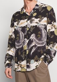 Koszula zapinana na guziki z ciemnym tłem i dużymi wzorami ptaków i kwiatów w odcieniach zieleni, bieli i niebieskiego; gładka faktura materiału.