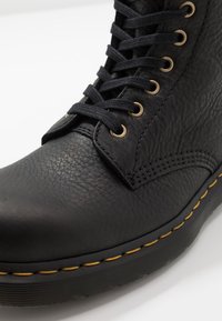 Stivaletto in pelle nera con superficie texturizzata, occhielli dorati, lacci neri e cuciture gialle a contrasto lungo la suola. Design a profilo basso.