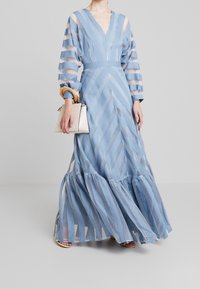 Ljusblå lång klänning med genomskinliga randiga ärmar, V-ringning och lager av volanger i nederkant. Matchad med en liten vit handväska.