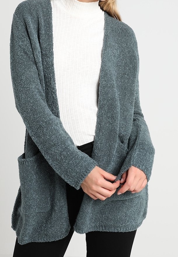 Cardigan bleu sarcelle doux et texturé avec une coupe décontractée, devant ouvert, deux poches avant, et manches longues, porté sur un col roulé côtelé blanc.