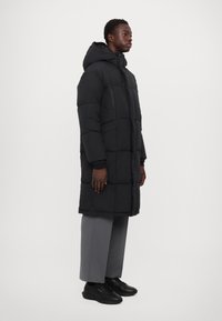 Abrigo de plumas negro con capucha, con secciones acolchadas, cierre frontal con cremallera y bolsillos laterales. Lleva puesto sobre pantalones grises y zapatos negros.