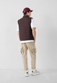 Bruine pufferbodywarmer over een wit t-shirt, gecombineerd met beige cargobroeken. Grijze sneakers met witte zolen; een bordeauxrode pet maakt de look compleet.