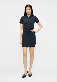 Jeune femme debout, main sur la hanche, portant une robe polo à manches courtes bleu marine à pois et des talons pointus noirs.