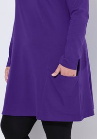 Lila Tunika-Kleid aus weichem Baumwollmaterial, mit langen Ärmeln, abgerundetem Saum und zwei seitlichen Taschen. Die Struktur wirkt glatt und leicht.