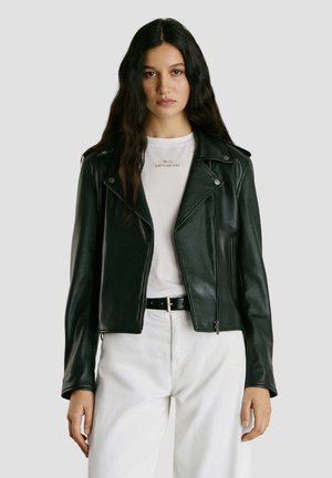 Jonge vrouw met lang donker haar, gekleed in een zwart leren jack over een witte T-shirt met de tekst "Notting Hill" en witte broek met een zwarte riem.