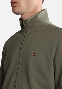 Uomo che indossa una giacca zip-up verde oliva con colletto alto e una piccola bandiera norvegese ricamata sul petto.