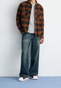 Camicia a quadri marrone e nera, t-shirt grigia, jeans larghi sbiaditi, sneakers nere e rosse con suola bianca. Sovrapposizione casual con tasche.