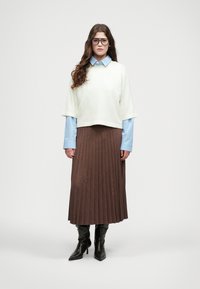 White cropped sweater over a blue striped shirt, paired with a brown pleated skirt and black knee-high boots. Glasses as an accessory.