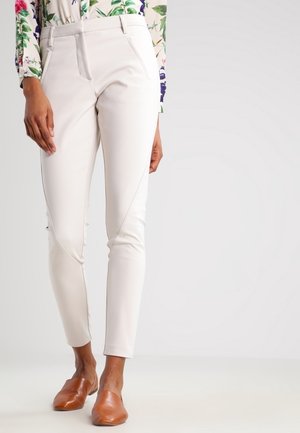 Kobieta ubrana w dopasowane białe spodnie, kwiecistą bluzkę z długim rękawem oraz brązowe skórzane wsuwane buty, stojąca na jednolitym tle.