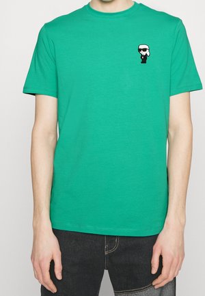 T-shirt en coton vert avec un col rond et des manches courtes, présentant un petit motif graphique noir et blanc sur la poitrine gauche.