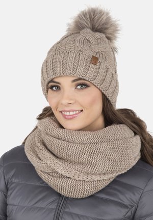 Beanie lavorato a maglia beige con un pompon in pelliccia sintetica e un piccolo tag in pelle, abbinato a una sciarpa infinita lavorata a maglia coordinata. Morbida texture, design a treccia.