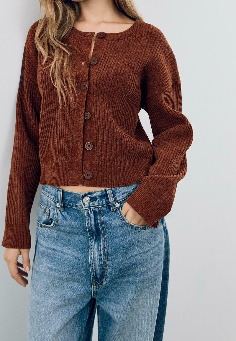 Femme portant un gilet en maille côtelée marron avec des boutons et un jean taille haute bleu, une main dans la poche, sur un fond uni.