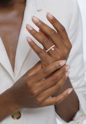 Donkere handen met natuurlijke nagels die een fijn parelringetje met een kleine bedel dragen, gekruist over een witte blazer.