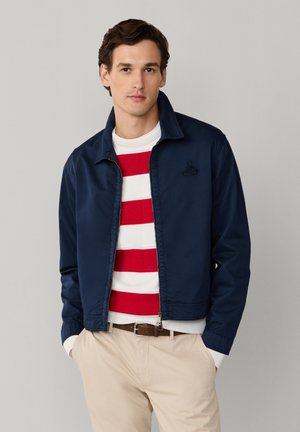 Hombre con chaqueta azul marino sobre suéter a rayas rojas y blancas, pantalones beige y cinturón marrón, sobre fondo gris liso.