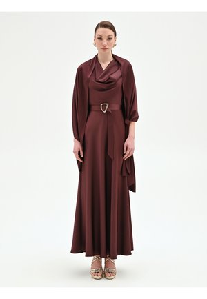 Femme portant une robe en satin bordeaux foncé avec un décolleté drapé, un châle assorti, une taille ceinturée et des talons dorés à lanières, debout sur un fond blanc uni.