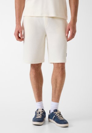 Homme portant un short texturé blanc cassé, des chaussettes blanches et des baskets bleu marine avec des lacets beiges, sur un fond uni.