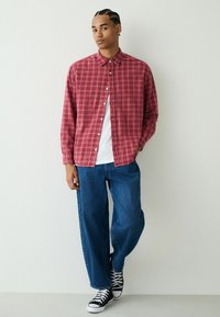 Rot-weiß kariertes Hemd mit Knopfleiste, getragen über einem weißen T-Shirt, kombiniert mit lockeren blauen Jeans und schwarzen High-Top-Sneakern.