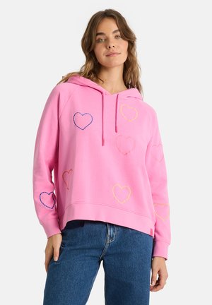 Frau trägt einen rosa Hoodie mit mehrfarbig gestickten Herzumrissen und blaue Jeans, steht vor einem weißen Hintergrund.