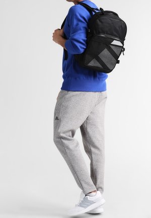 Personne portant un pantalon de survêtement Adidas gris, des baskets blanches, un sweat bleu, et portant un sac à dos noir sur une épaule, debout devant un fond blanc.