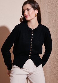 Svart stickad cardigan med rund halsringning, guldknappsstängning och sprucken nederkant. Slät textur och åtsittande ärmar.