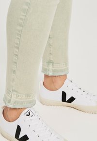 Vaqueros verde claro con textura desvanecida y bajos deshilachados, combinados con zapatillas blancas que presentan un logo negro prominente.