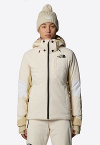Giacca imbottita beige con zip nera e accenti delle tasche, con pannelli bianchi sulle braccia e colletto alto. Berretto lavorato a maglia color crema con pom-pom.